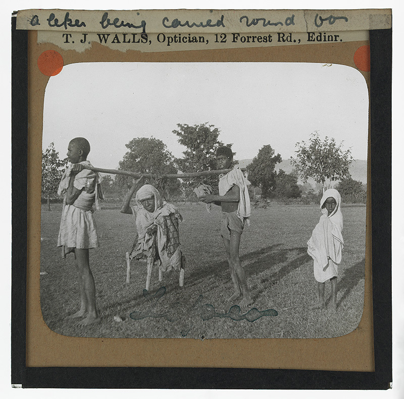 'A Leper'. Photograph of a leper being carried by two boys with a pole and hanging bench 'for alms' in Keonjhar, Orissa, India in the late 19th or early 20th century.