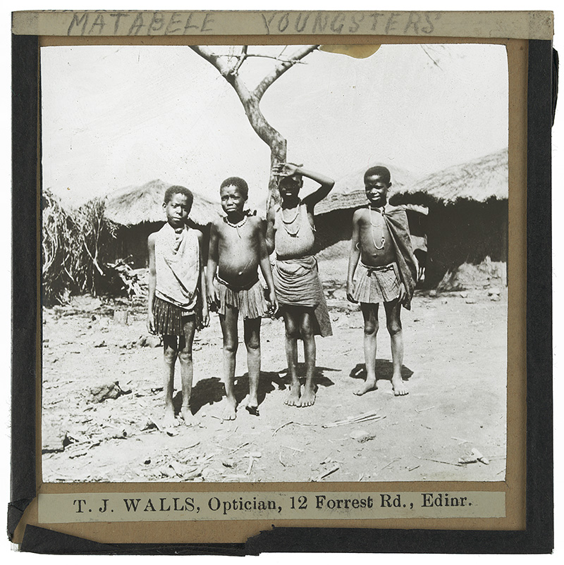 'Matabele Youngsters'. Photograph of four Matabele [Ndebele] children in traditional dress standing in front of a tree and some huts in the early 20th century. 
