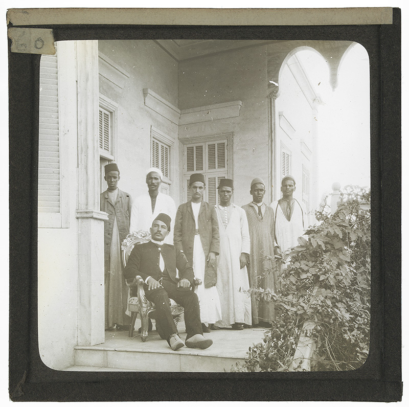 'Group of Egyptian Men on a Porch'. Photograph of a group of Egyptian men on the porch of a country house in Egypt in the late 19th or early 20th century. One man sits in an ornate chair while six other men stand behind him. 