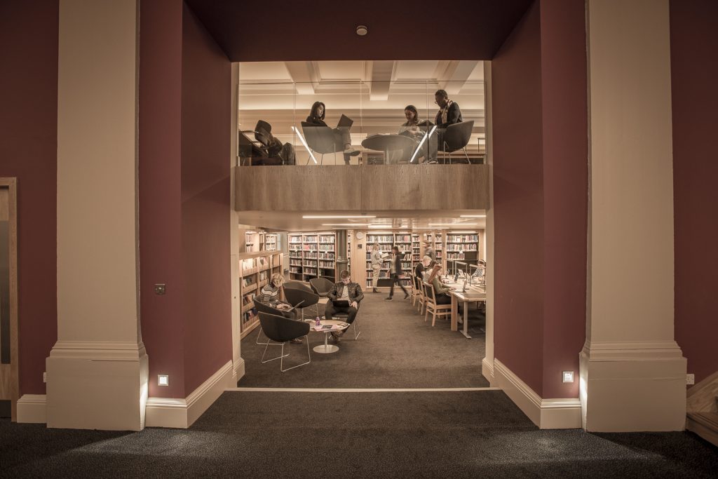 Students sit in armchairs on the mezzanine level, visible in the top half of an archway. Students browse books in the library stacks on the ground floor. 