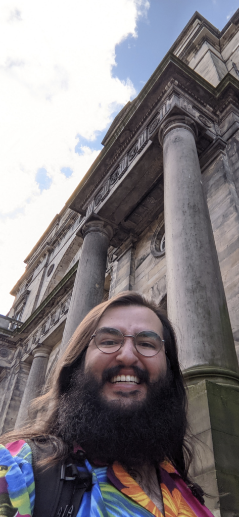Noah stands in the foreground of the picture, smiling at the camera. He has dark hair and beard, and is wearing glasses. He has a colourful tshirt on. Behind him the pillars of a building in Old College are visible. The photo is taken with the camera from a low angle so that a portion of blue sky and white clouds are also visible.