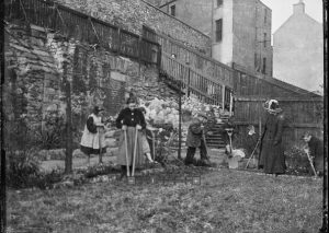 Black and white print from a glass plate negative showing, four children and a woman working in the Children's Garden, Johnston Terrace, Edinburgh (Patrick Geddes Collection, Ref: Coll-1167/B/27/10/9)