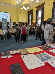 A leaflet display on a table, with other stands around the room and small groups of students stood in between them.