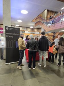 People gathered at a welcome desk inside the University of Edinburgh Main Library with stairs and modern interior in the background.