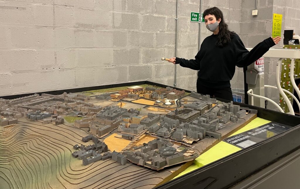 A woman wearing a facemask and all black stands beside the model of the city with a tool in her hand. 