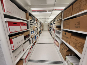 Colour photograph within a strongroom. The floor, shelving and ceiling are white, and the lighting is fluorescent. The shelving rows run down the left and right of the image, and on the shelves are cardboard boxes, lever arch files, loose papers and assorted documents. 