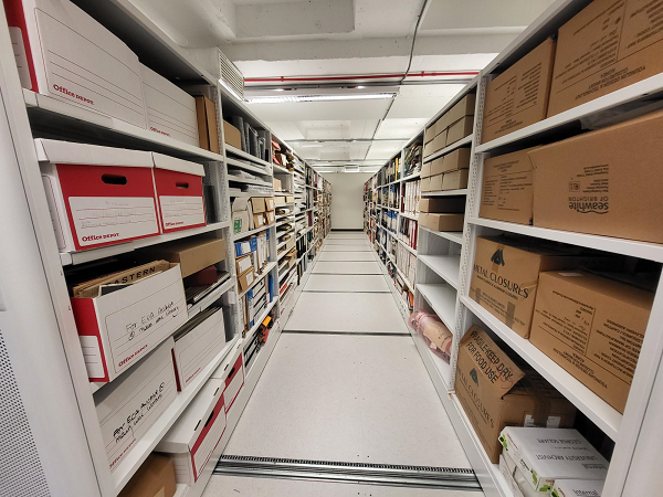 Colour photograph within a strongroom. The floor, shelving and ceiling are white, and the lighting is fluorescent. The shelving rows run down the left and right of the image, and on the shelves are cardboard boxes, lever arch files, loose papers and assorted documents.