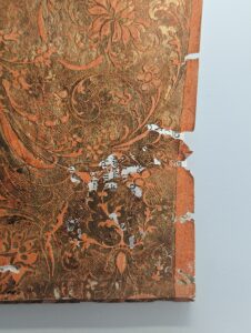 This image shows the corner of a cover of a paper bound volume. The cover is decorated in swirling designs in orange and gold. There are some holes in the cover, revealing the page below. 