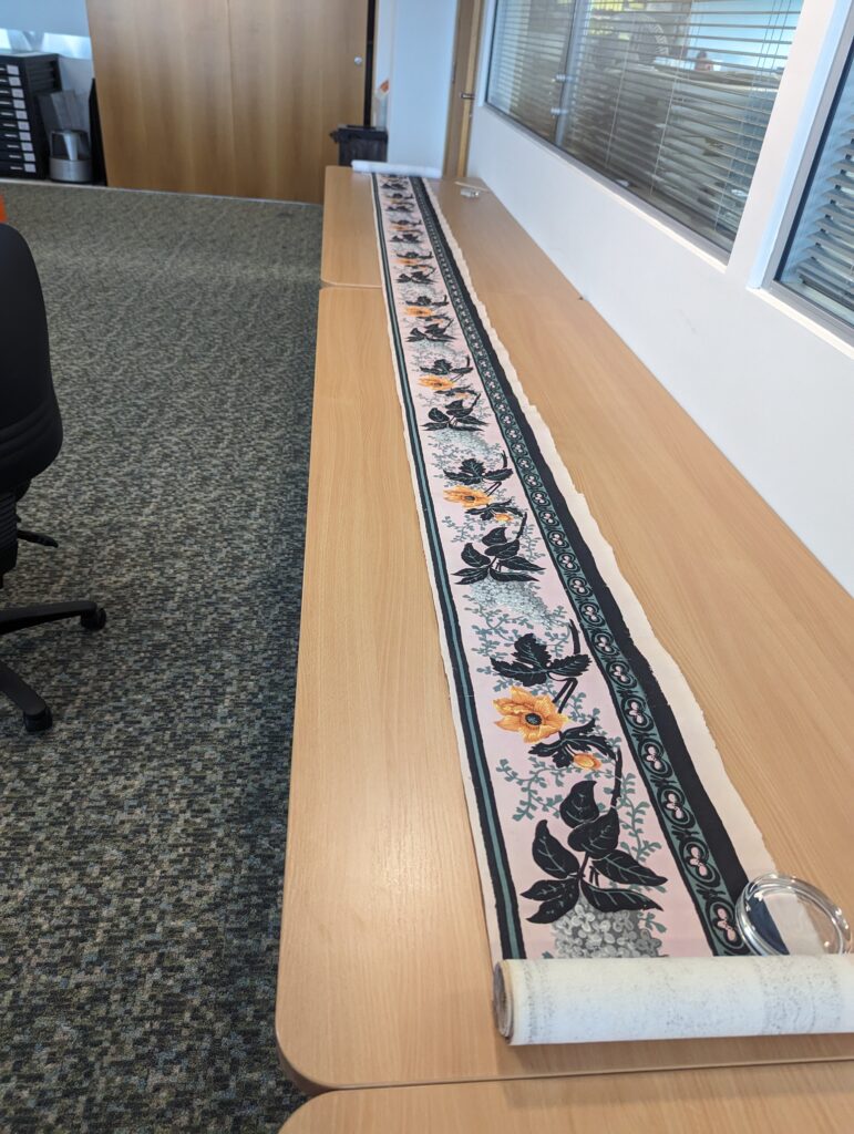 A long, narrow, sheet of wallpaper has been unrolled across a long table. It has a floral design with blue and orange colours, and a thick blue boarder. 