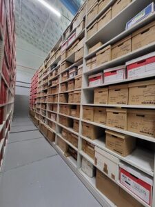 Colour image of white shelving along a long grey floor corridor. Three or four bankers boxes sits on each shelf, showing an extensive amount of material.