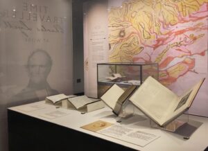 An exhibition display case containing two handwritten notebooks and two printed books. We cannot see the contents, but they are displayed open for visitors to read. The wall in the background is covered with a map of the Strathmore Valley, hand painted in yellow, pink and orange watercolour.