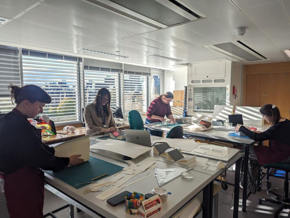 Four people are gathered round a large table in a busy conservation studio. They are working with their hands and lots of materials and kit are out across the table. 