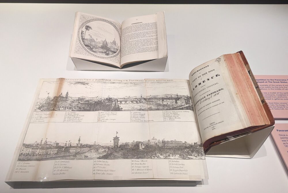 Two books are pictured open in a display case. One has a long fold out picture of a panorama. They are both mounted on white box board book cradles.