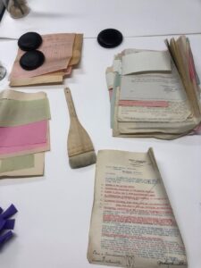 A white table with an arrangement of grimy papers and a brush used for cleaning placed at an angle. Some black circular weights holding the papers down. 