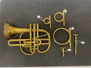 Brass instrument resting on a grey flat surface, oriented with the bell to the left. On the right of the image, five sections or parts of these instrument arranged 