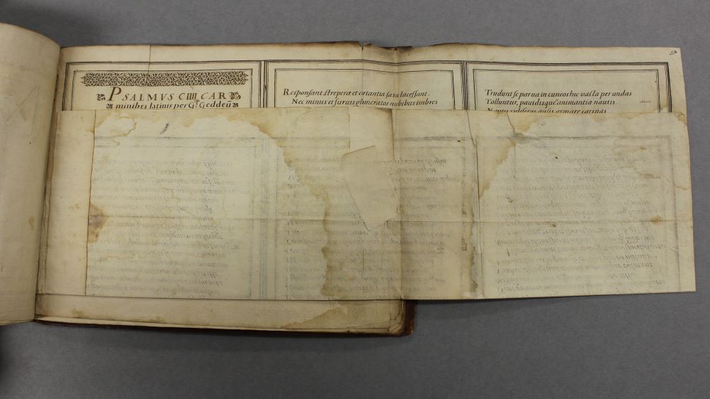 An open book with one oversized page partially folded over on itself. The paper has brown water stains and old tears have been with mended with paper patches stuck on the back.