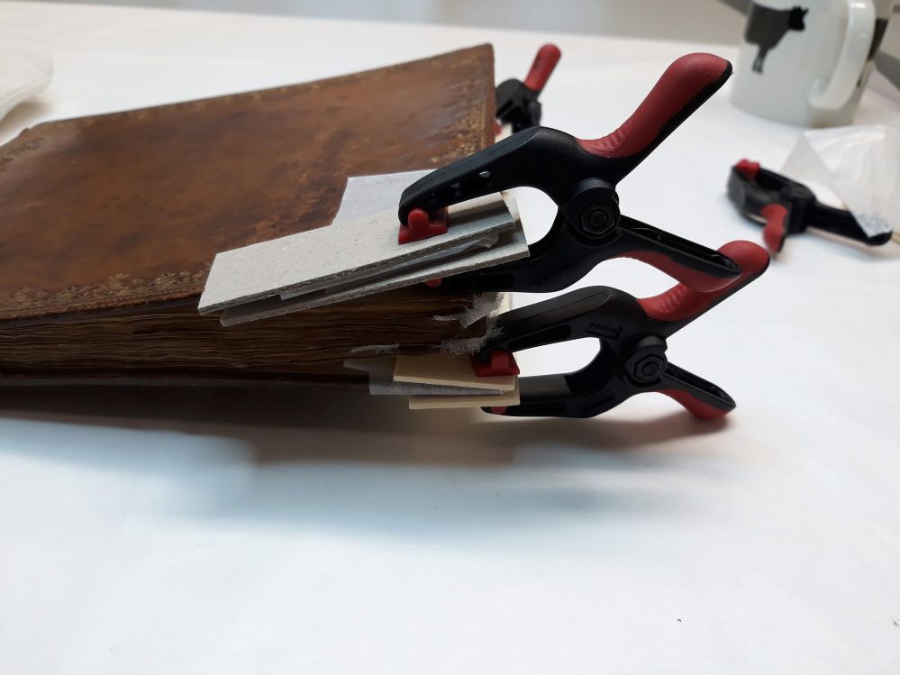 A brown leather book. Each of the four corners of its covers are clamped between pieces of paper board.