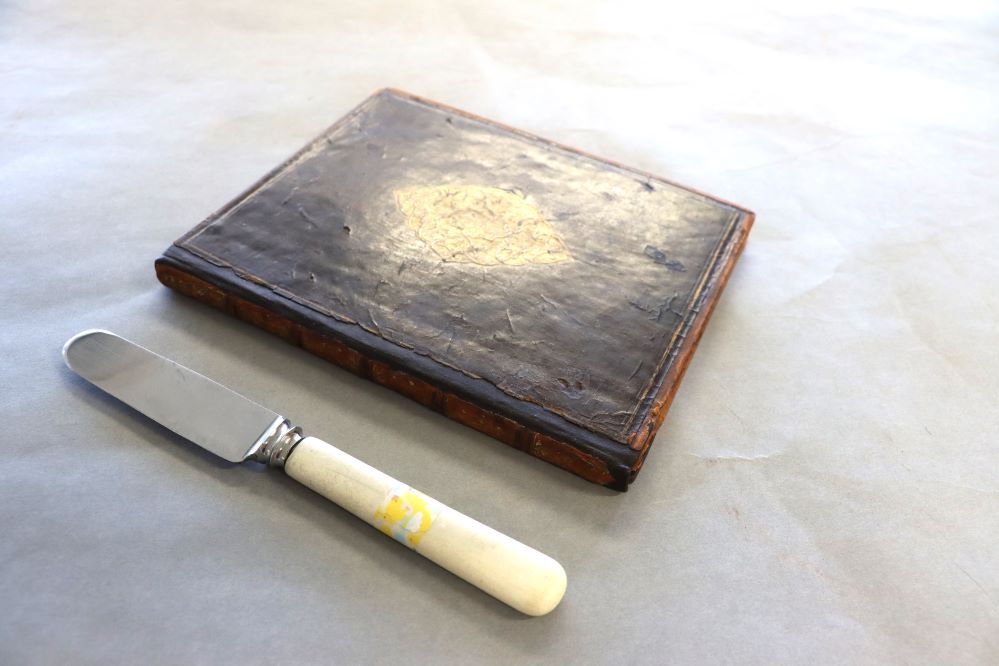 A white-handled knife next to a brown leather book with gold decoration. The front cover of the book is attached and there is a strip of new dark brown leather between the spine and the front cover. 