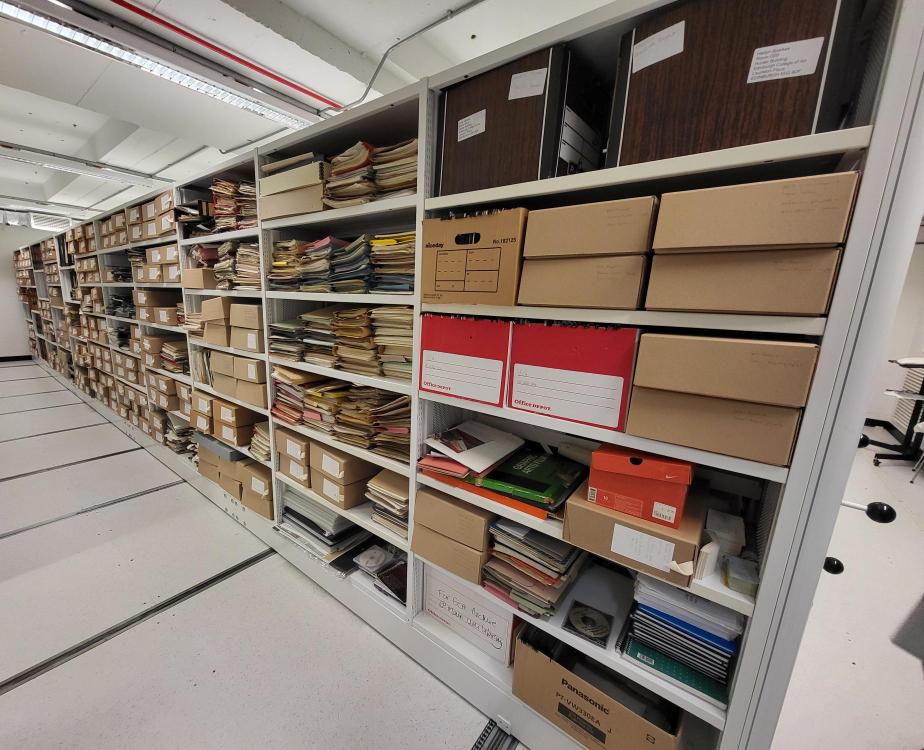 Colour photograph of white metal shelving. Boxes of various sizes, colour and condition house material, and there are many loose folders and items on the shelves as well.