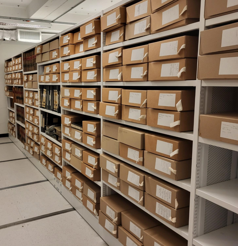 Colour photograph of white metal shelving. On the shelves are brown archival boxes, six to a shelf in three columns of two. Loose lever arch files or bound volumes are visible on some shelves.