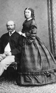 Full-length black-and-white portrait of a young woman wearing a fashionable gown with pagoda sleeves, her hair styled up in ringlets, standing beside her seated husband.