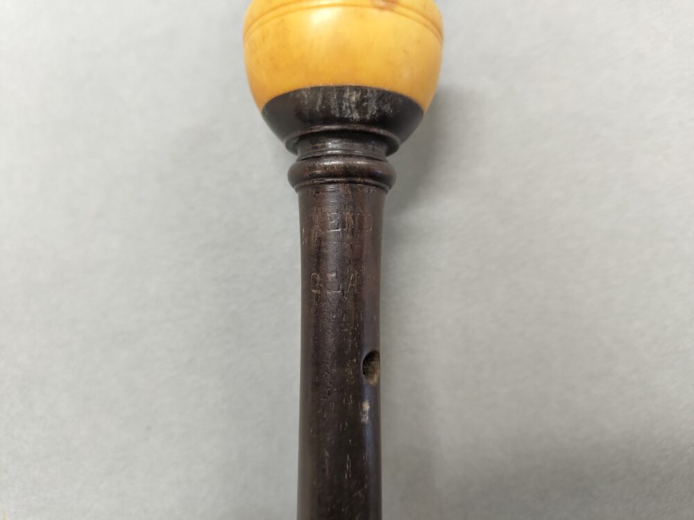 Upper section of a bagpipe chanter. The body is made of dark wood, conical, and has a stamp partially visible, the discernible writing reads “ENDER” and on a second line below “GLAS”. The chanter ends in a chalice shape where the black wood body meets an ivory ferrule. 