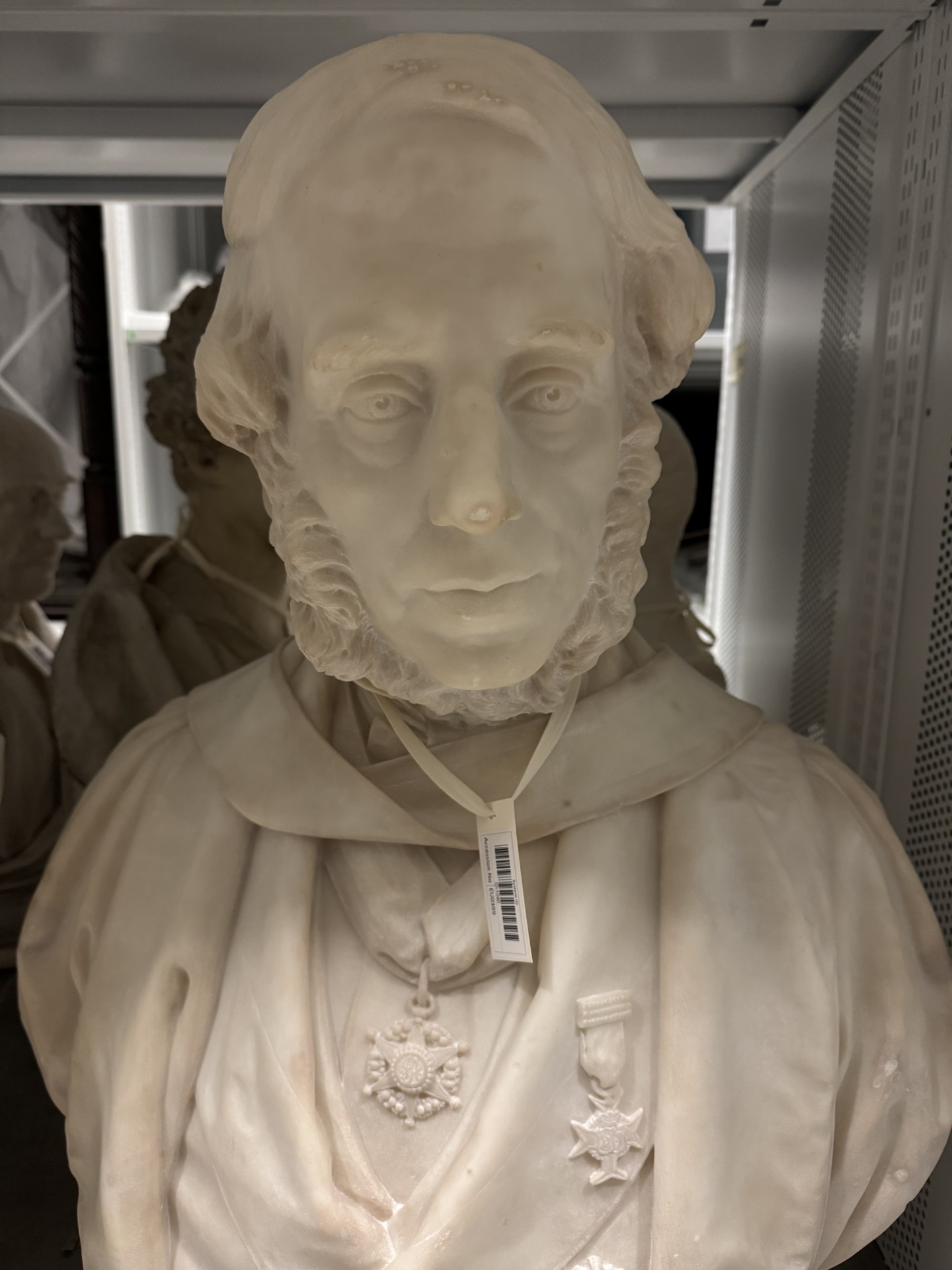 Image of a white marble bust of an elder man with a balding pate and mutton chops. A barcode label on a white cotton ribbon is tied around his neck.