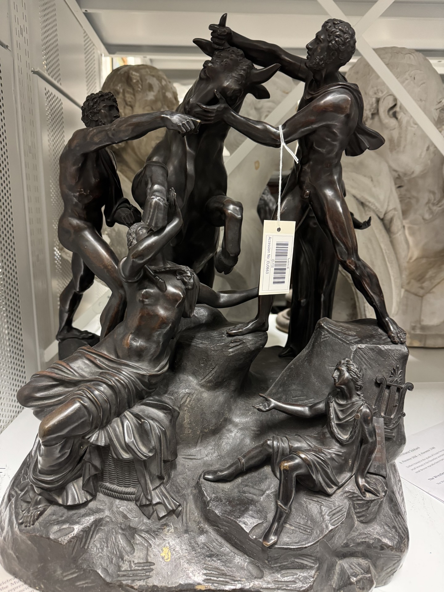 Bronze sculpture with male and female figures wrestling a bull. A barcode label is hanging off of the right hand figure's arm.