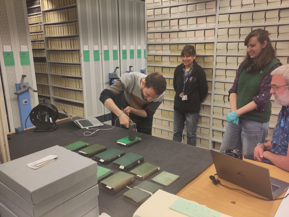 A scientist points a grey hand-held scanning tool at a bright green book resting on a table. A red light is illuminated on the top of the tool.
