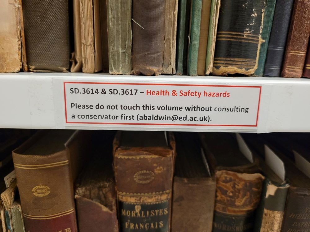 A row of books sitting on a shelf. A label on the shelf states that two of the books are hazards and should not be handled without consulting a conservator.
