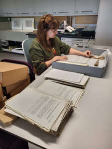 A woman sits at a grey table. She is holding a small beige sponge and is brushing at pages stained with soot and dirt. The pages are resting in a card enclosure to prevent the table from getting dirt from the soot particles. There is also two brushes, her laptop, and a pile of cleaned pages on the table.