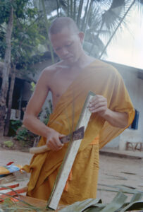 A monk in orange robes cutting palm leaves using a sharp knife and wooden template.