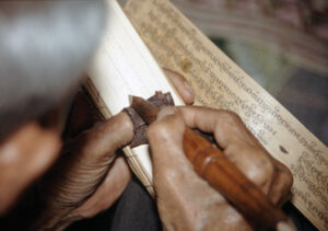 Hands holding a pointed stylus and a palm leaf. The hand appears to be inscribing text on to the leaf. 