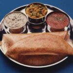 Photo of a folded dosa pancake on a tray with dishes of savoury sauces. 