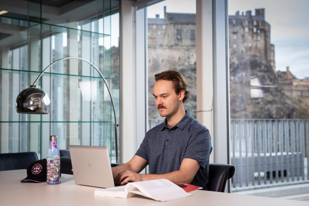 Young man with laptop on table with Edinburgh Castle seen through the window.