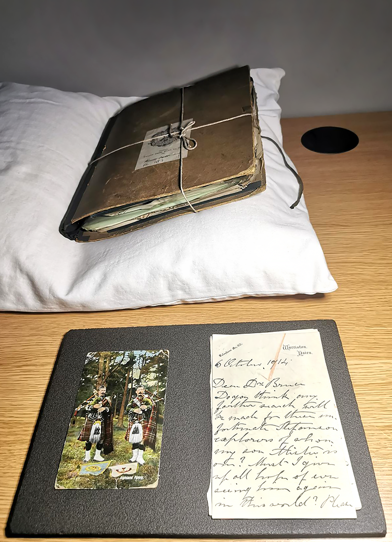 Archive materials laid out on a table. In the background is an old, worn looking book held together with string sits on a conservation-safe pillow used for safely supporting books with fragile bindings. In the foreground is a plastazote foam sheet which is also conservation safe, on top rests a postcard with the image of two Highland pipers in a forest, beside it is an example of one of the letters from the collection.
