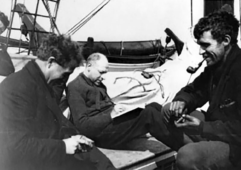 A black and white image of three men on a sailing ship. They are sat in a circle smiling and socialising, none are looking at the camera. The man on the right is smoking a pipe.