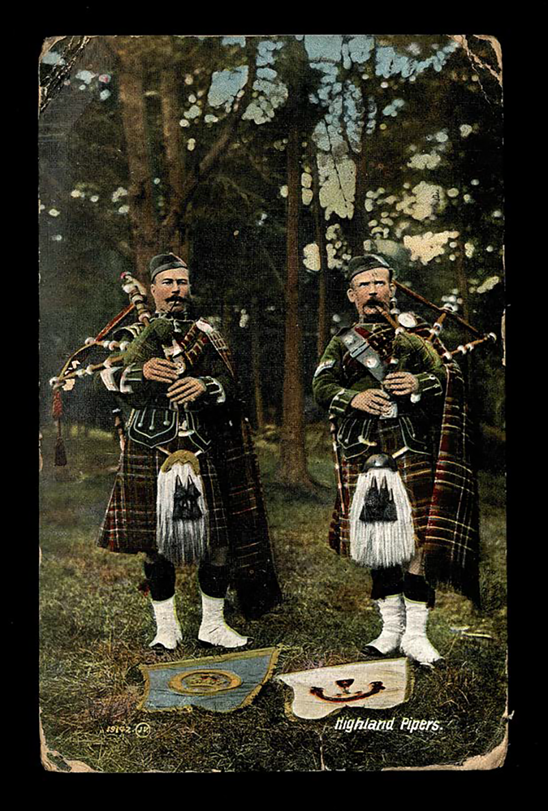 Postcard front image. Two highland pipers stood in a forest.