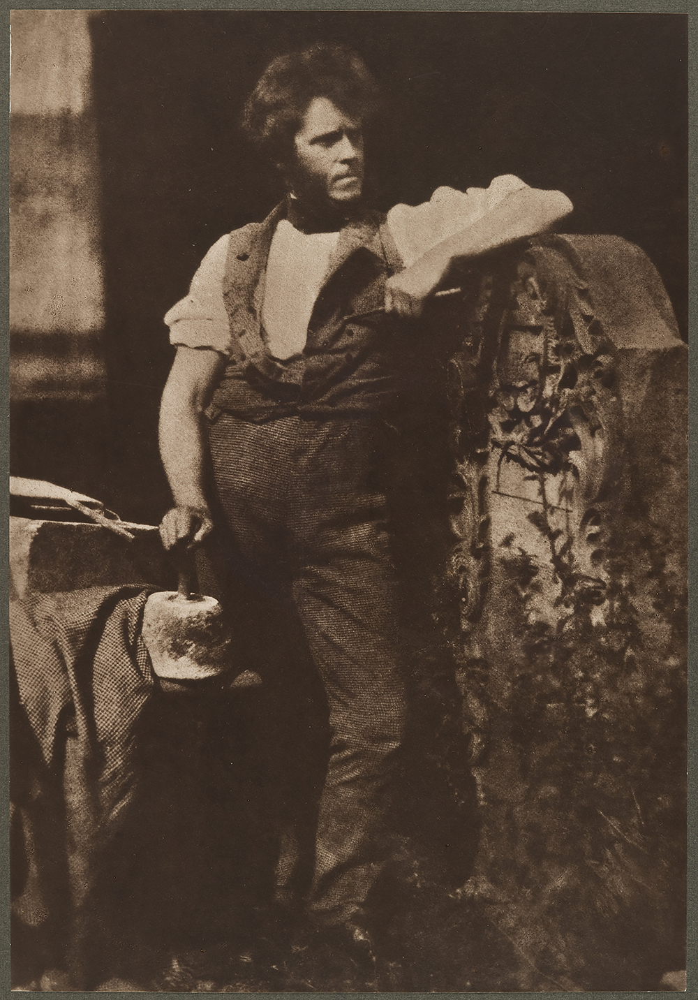 Aged, sepia photograph of a man in victorian dress, posed leaning on a gravestone, his other hand resting on stonemasonry equipment and tools.