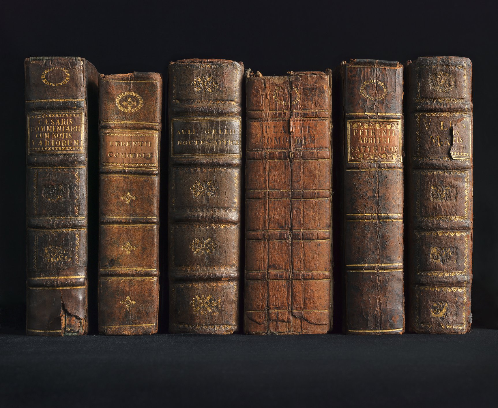 Row of books, bound in old, cracked brown leather resting on a black background with spines facing outwards.