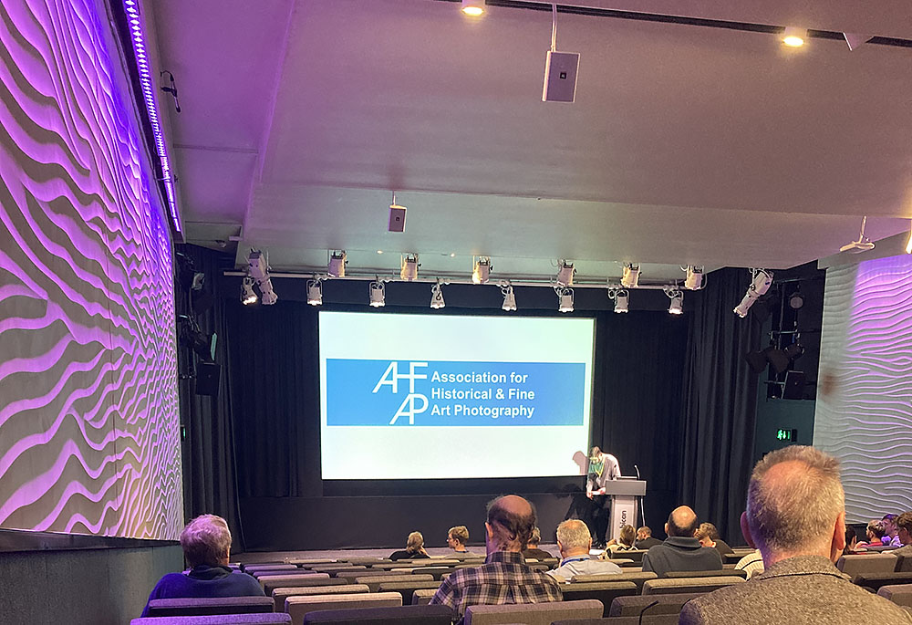 Image taken in a lecture theatre in the Barbican Centre. Taken from the perspective of the audience looking towards a screen with the AHFAP logo projected on it.