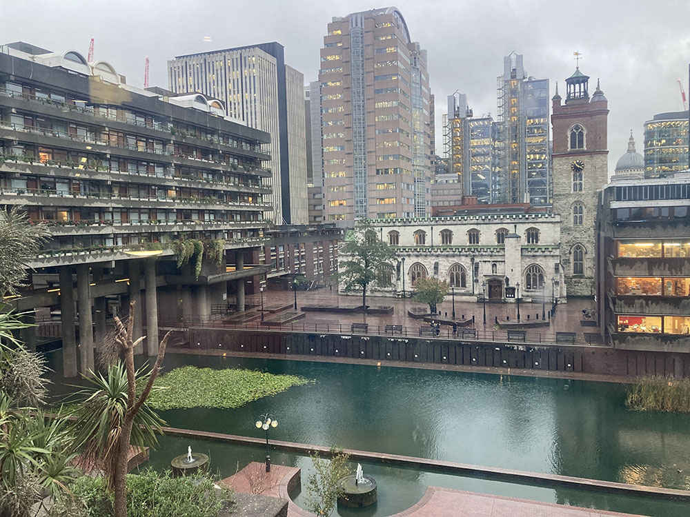 Image taken looking out from within the Barbican Centre on a wet day. There is a square, green coloured pond surrounded by several buildings of varying heights. 