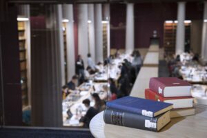 Image of books stacked on a small table in the foreground on the mezzanine of the Law Library, looking out across a room full of students studying in the Senate Room (out of focus).