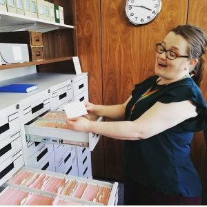 Louise is standing in side profile in a card index. She is showing a card to the camera