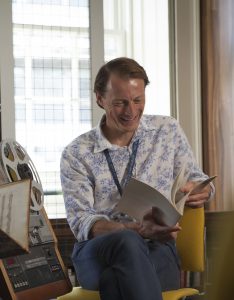 Stuart sits to the right of a reel to reel machine. He is holding a booklet open and looking at it