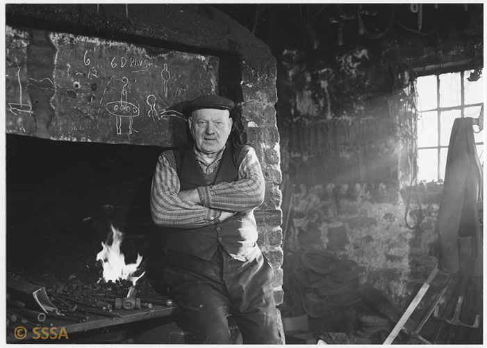 Black and white image of a blacksmith who is sitting on the edge of a fireplace, a flame behind him. TThere are chalk drawings on the breast of the chimney. Light is streaming in a window just out of shot