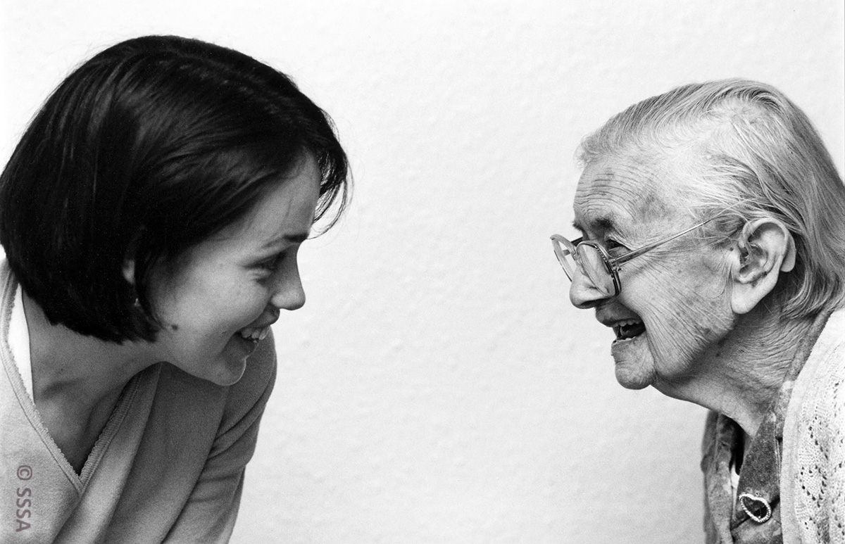 Black and white image of two women in side profile, Dr K Campbell looks at her interviewee Lizzie Angus. They smile openly towards eachother