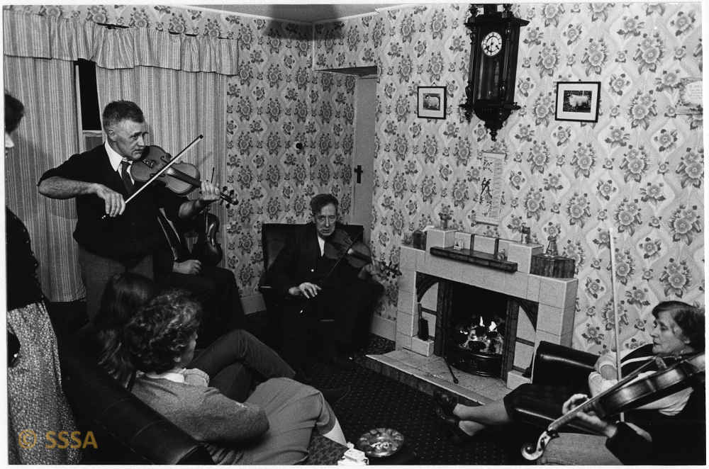 black and white image of a group of people in a sitting roo,. a man stands with a fiddle , two others are seated with fiddles. 