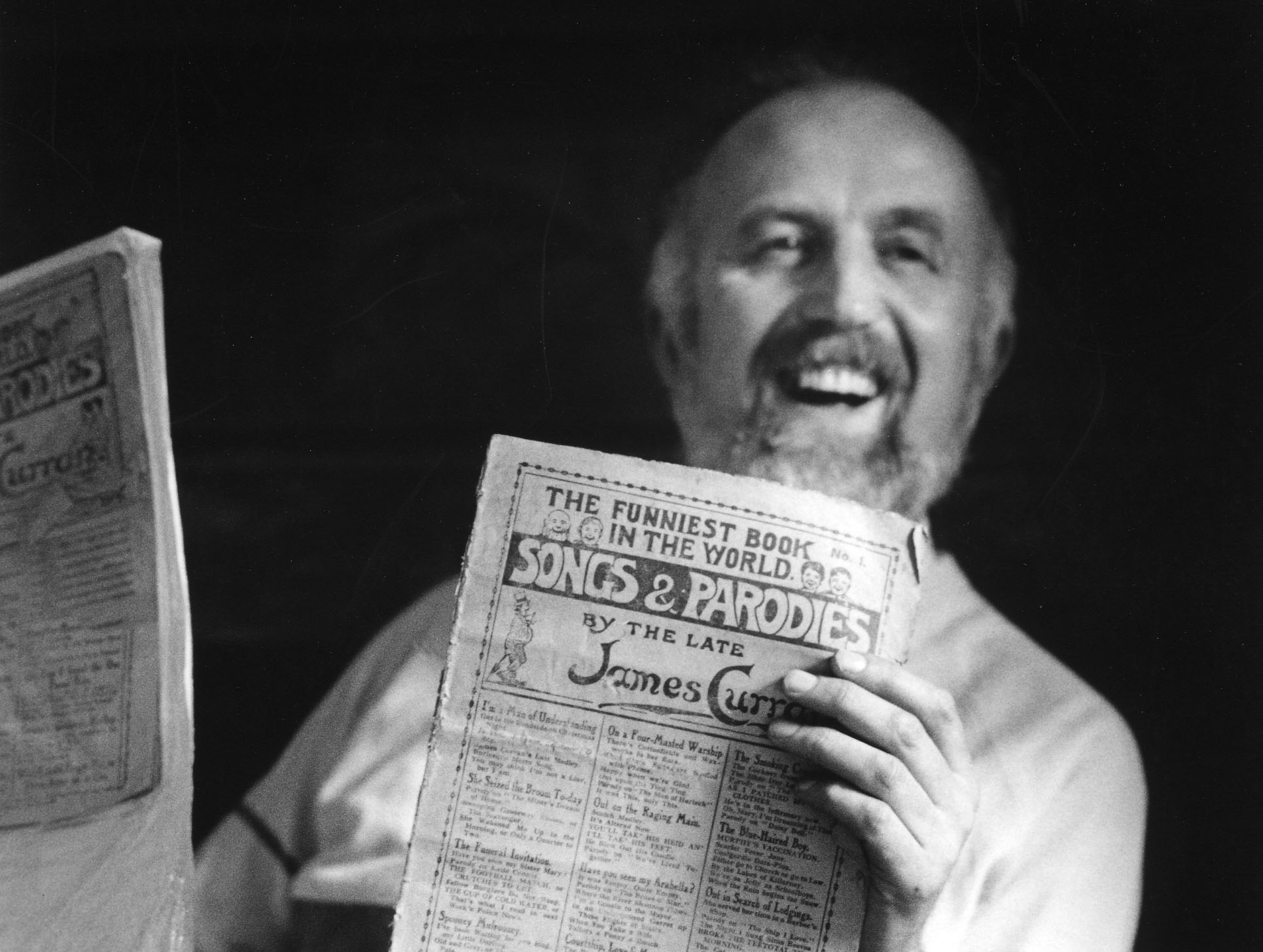 black and white image of adam sitting back in a chair in each hand he holds a newspaper style pamphlet. He is smiling broadly