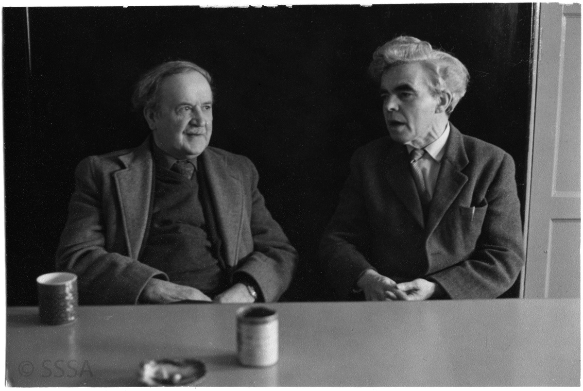 a black and white image of two men sitting at a table.  They are turned into one another in conversation